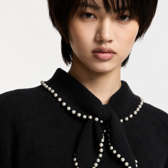 A close-up portrait of a model wearing a black knit sweater with a high collar decorated with a bow and a border of white pearls.