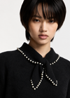 A close-up portrait of a model wearing a black knit sweater with a high collar decorated with a bow and a border of white pearls.