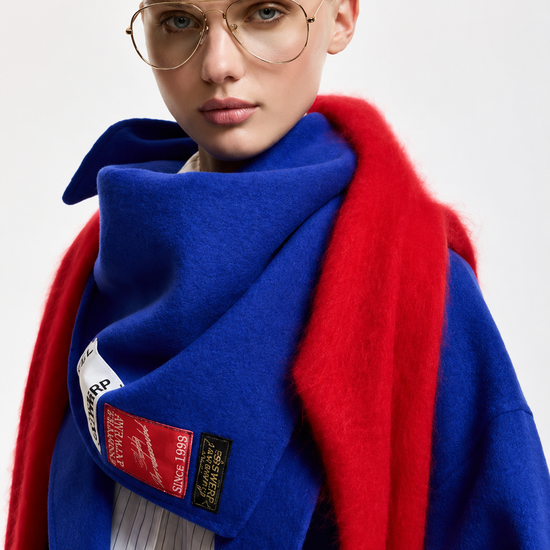 A close-up of a model wearing a vibrant royal blue coat with a contrasting bright red shawl, a thick blue scarf with a red patch, and round gold-framed glasses against a light gray background.