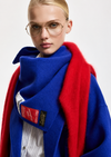 A close-up of a model wearing a vibrant royal blue coat with a contrasting bright red shawl, a thick blue scarf with a red patch, and round gold-framed glasses against a light gray background.