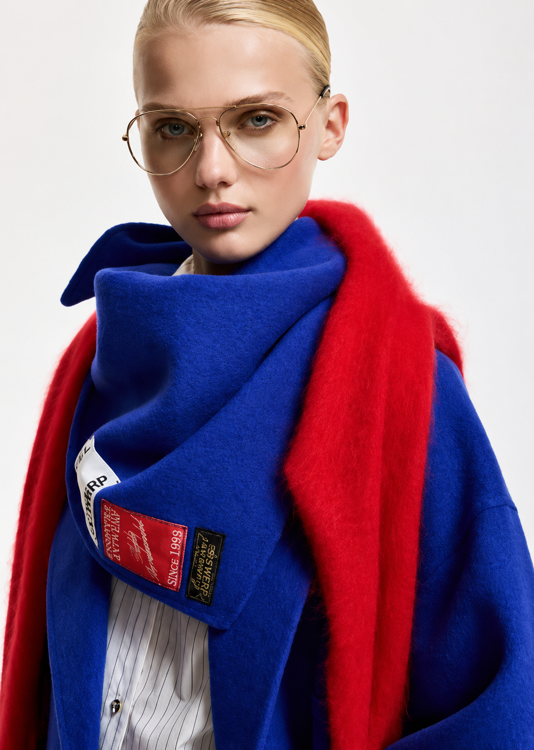 A close-up of a model wearing a vibrant royal blue coat with a contrasting bright red shawl, a thick blue scarf with a red patch, and round gold-framed glasses against a light gray background.