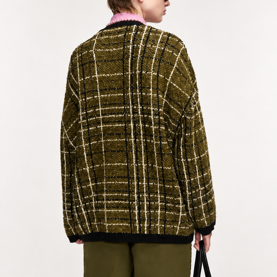 A model shown from the back wearing an oversized tweed plaid cardigan in olive green and beige, paired with olive green trousers and black sunglasses, holding a black handbag against a plain white background.