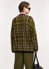 A model shown from the back wearing an oversized tweed plaid cardigan in olive green and beige, paired with olive green trousers and black sunglasses, holding a black handbag against a plain white background.