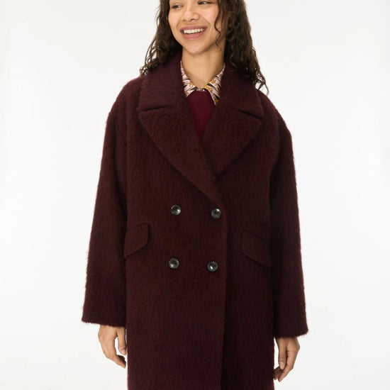 A woman wearing a dark burgundy double-breasted fuzzy coat over a patterned shirt and mauve pants, standing against a plain white studio background.