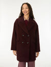 A woman wearing a dark burgundy double-breasted fuzzy coat over a patterned shirt and mauve pants, standing against a plain white studio background.