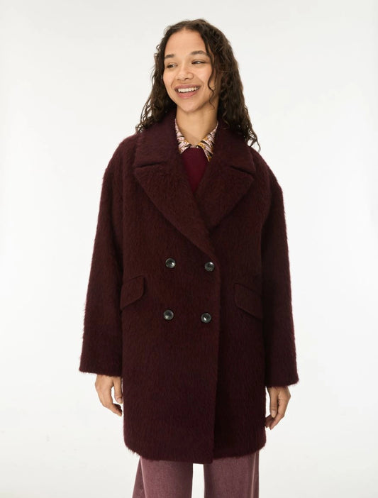 A woman wearing a dark burgundy double-breasted fuzzy coat over a patterned shirt and mauve pants, standing against a plain white studio background.