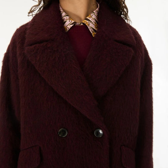 A close-up of a model wearing a dark burgundy fuzzy double-breasted coat over a burgundy top and a patterned collared shirt.