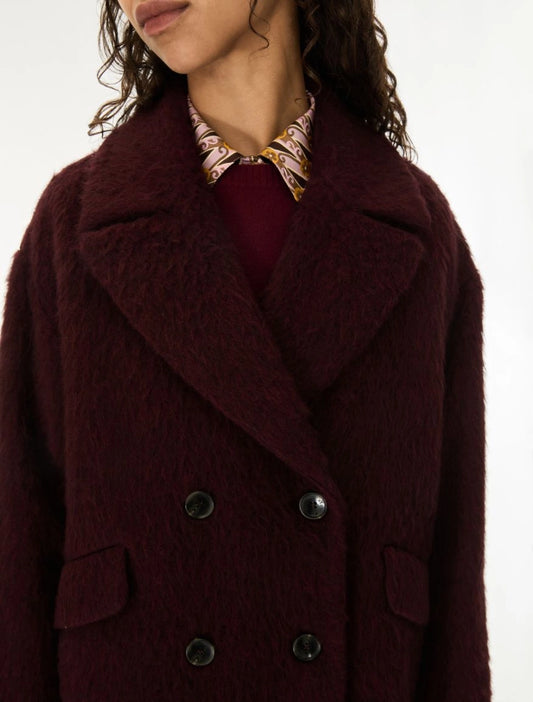 A close-up of a model wearing a dark burgundy fuzzy double-breasted coat over a burgundy top and a patterned collared shirt.