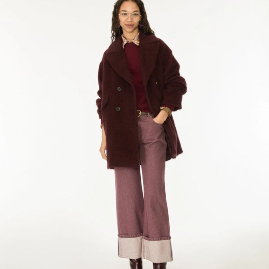 A model stands against a plain white studio background wearing a dark burgundy fuzzy double-breasted coat over a burgundy top, paired with dusty mauve wide-leg jeans featuring light beige turned-up cuffs and dark brown boots.