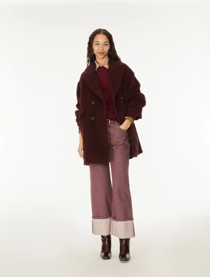 A model stands against a plain white studio background wearing a dark burgundy fuzzy double-breasted coat over a burgundy top, paired with dusty mauve wide-leg jeans featuring light beige turned-up cuffs and dark brown boots.