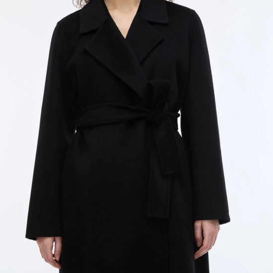 woman wearing A black long coat with a tie belt, shown on a plain light gray background. close up
