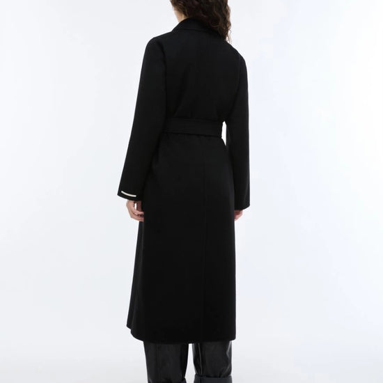woman wearing a black long coat with a tie belt, shown on a plain light gray background rear view