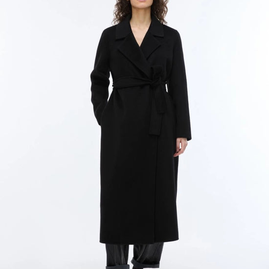 A woman modeling a long black wrap coat with a self-tie belt, paired with dark trousers and fuzzy black shoes, against a plain white studio background.