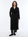 A woman modeling a long black wrap coat with a self-tie belt, paired with dark trousers and fuzzy black shoes, against a plain white studio background.