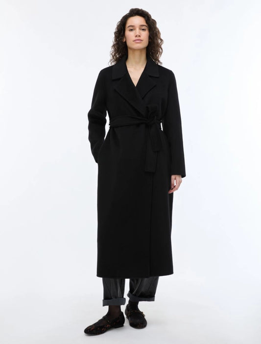 A woman modeling a long black wrap coat with a self-tie belt, paired with dark trousers and fuzzy black shoes, against a plain white studio background.