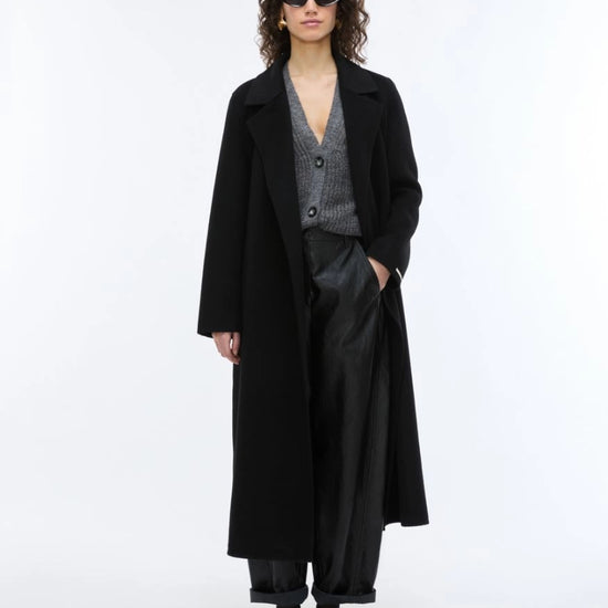 A model stands against a plain white studio background wearing a long black coat over a gray patterned V-neck sweater, black wide-leg trousers, and black shoes.