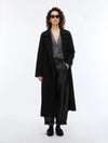 A model stands against a plain white studio background wearing a long black coat over a gray patterned V-neck sweater, black wide-leg trousers, and black shoes.
