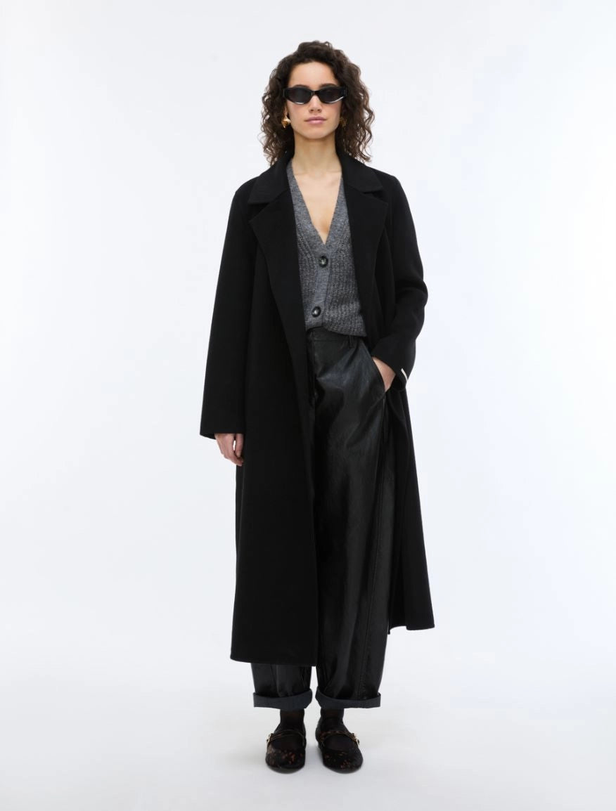A model stands against a plain white studio background wearing a long black coat over a gray patterned V-neck sweater, black wide-leg trousers, and black shoes.