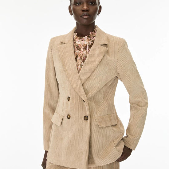 A model wearing a beige corduroy double-breasted blazer with matching trousers and a patterned blouse, photographed against a plain white studio background.