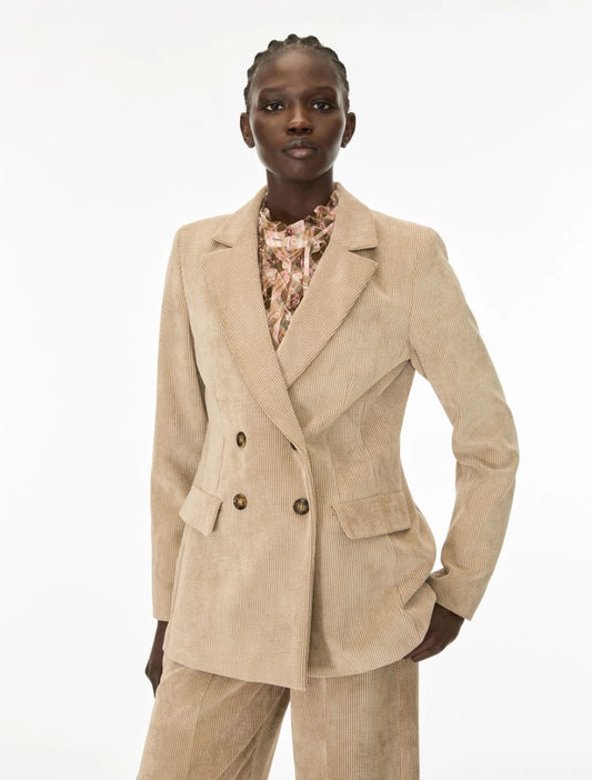 A model wearing a beige corduroy double-breasted blazer with matching trousers and a patterned blouse, photographed against a plain white studio background.