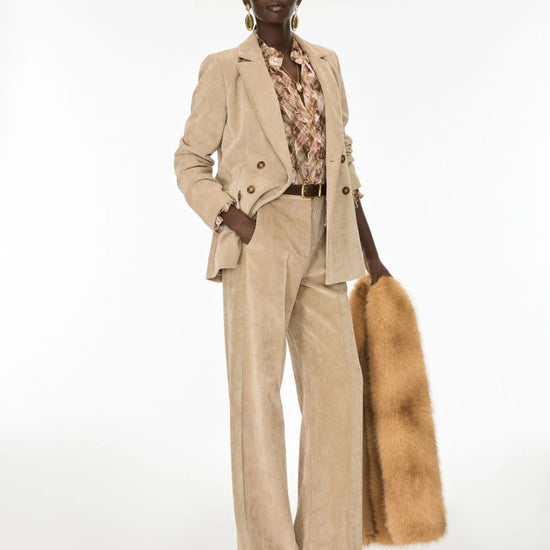 A model stands against a white studio background wearing a beige corduroy blazer and matching wide-leg trousers, layered over a floral shirt, and holding a tan fur stole.