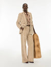 A model stands against a white studio background wearing a beige corduroy blazer and matching wide-leg trousers, layered over a floral shirt, and holding a tan fur stole.