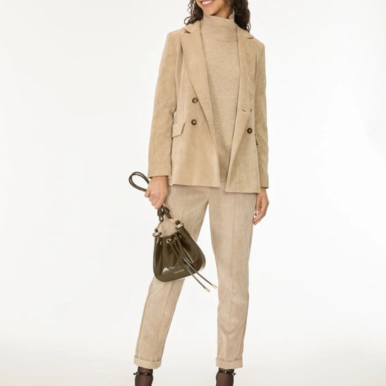 A woman modeling a beige double-breasted blazer, matching corduroy trousers, and a beige knit mock-neck top, accessorized with a dark brown bucket bag and black ankle boots against a white studio background.