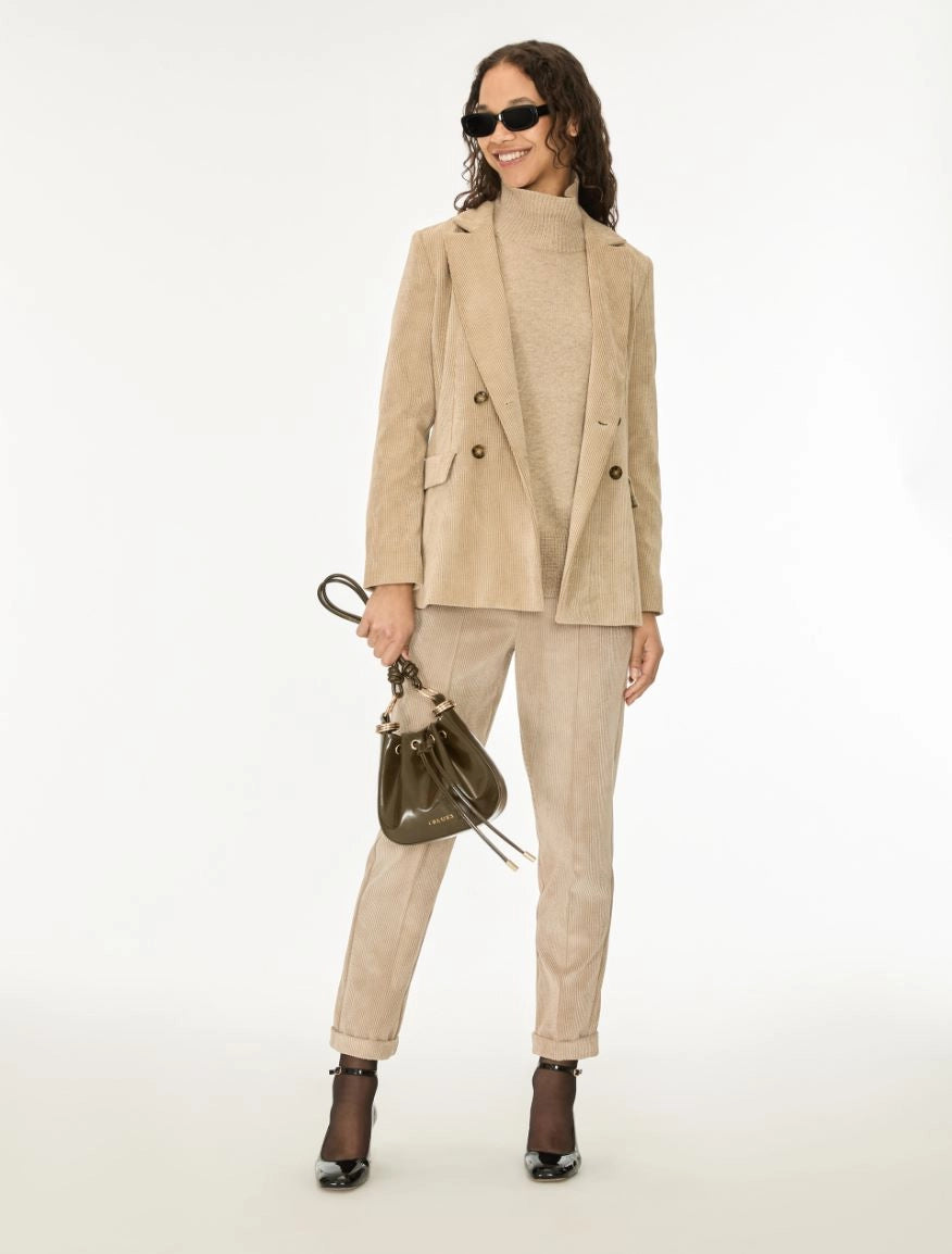 A woman modeling a beige double-breasted blazer, matching corduroy trousers, and a beige knit mock-neck top, accessorized with a dark brown bucket bag and black ankle boots against a white studio background.