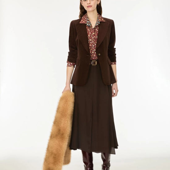 A model stands against a plain white studio background wearing a dark brown tailored blazer over a patterned collared shirt, a dark brown midi skirt, dark brown knee-high boots, and holding a tan fur stole.