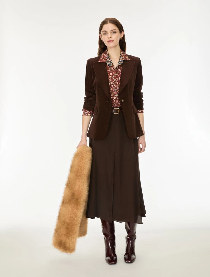 A model stands against a plain white studio background wearing a dark brown tailored blazer over a patterned collared shirt, a dark brown midi skirt, dark brown knee-high boots, and holding a tan fur stole.