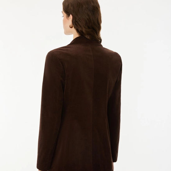 A woman viewed from the back wearing a dark brown suede blazer and a dark skirt against a plain white studio background.