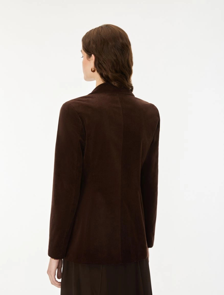 A woman viewed from the back wearing a dark brown suede blazer and a dark skirt against a plain white studio background.