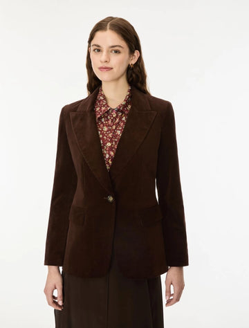A woman modeling a dark brown corduroy blazer over a patterned collared blouse and dark skirt, photographed against a clean white studio background.