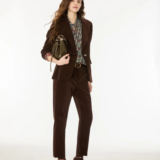 A woman models a dark brown corduroy pantsuit, featuring a tailored blazer and matching trousers, styled with a patterned blouse, brown leather ankle boots, and a dark green handbag against a plain white studio background.