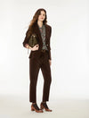 A woman models a dark brown corduroy pantsuit, featuring a tailored blazer and matching trousers, styled with a patterned blouse, brown leather ankle boots, and a dark green handbag against a plain white studio background.