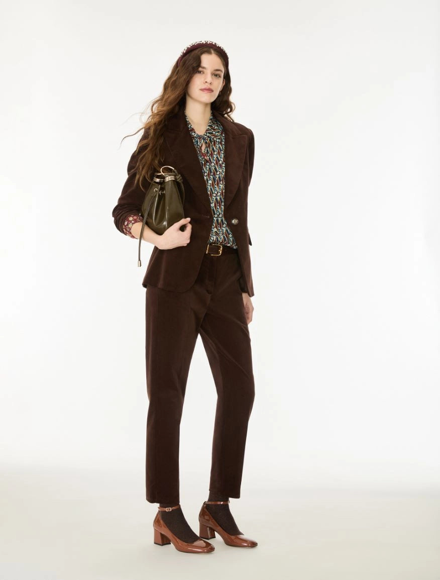 A woman models a dark brown corduroy pantsuit, featuring a tailored blazer and matching trousers, styled with a patterned blouse, brown leather ankle boots, and a dark green handbag against a plain white studio background.