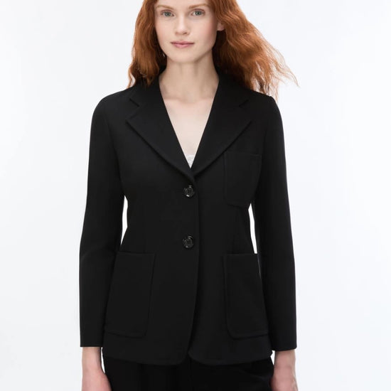 A woman with red hair wearing a black tailored blazer with notch lapels, two front buttons, and flap pockets, photographed against a clean white studio background.