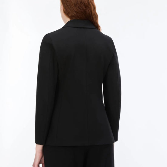 A woman with red hair is shown from the back wearing a black long-sleeve blazer and matching black trousers against a plain white studio background.
