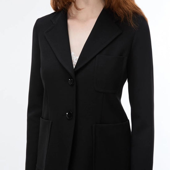 A woman wearing a tailored black blazer with notch lapels, two front buttons, flap pockets, and a structured silhouette, shown against a plain light background.