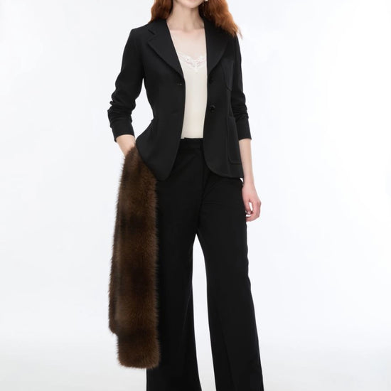 A woman modeling a black tailored blazer and matching wide-leg trousers, styled with a cream top and holding a dark brown fur stole against a plain white studio background.