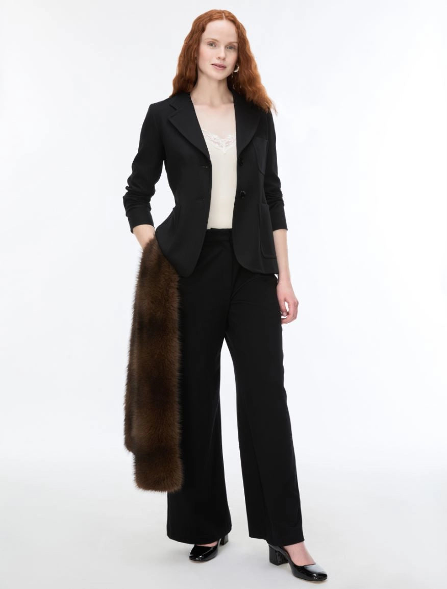 A woman modeling a black tailored blazer and matching wide-leg trousers, styled with a cream top and holding a dark brown fur stole against a plain white studio background.