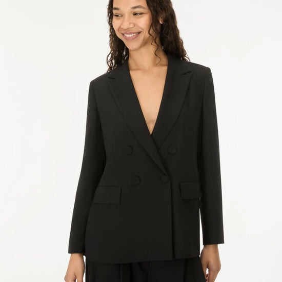 A woman wearing a black double-breasted blazer with a deep V-neckline and matching black trousers, standing against a plain white studio background.