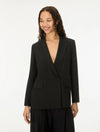 A woman wearing a black double-breasted blazer with a deep V-neckline and matching black trousers, standing against a plain white studio background.