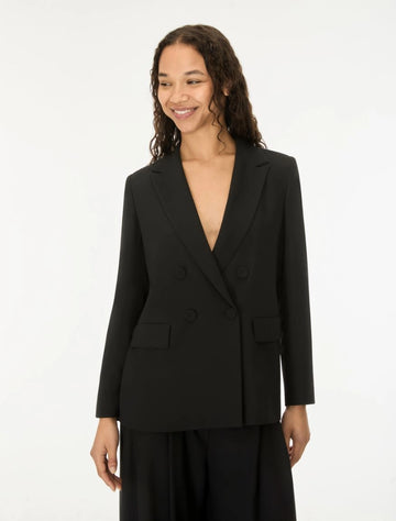 A woman wearing a black double-breasted blazer with a deep V-neckline and matching black trousers, standing against a plain white studio background.
