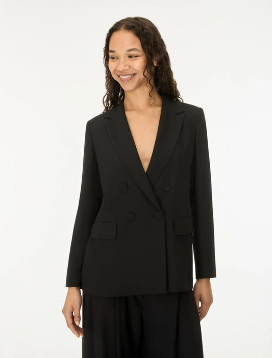 A woman wearing a black double-breasted blazer with a deep V-neckline and matching black trousers, standing against a plain white studio background.