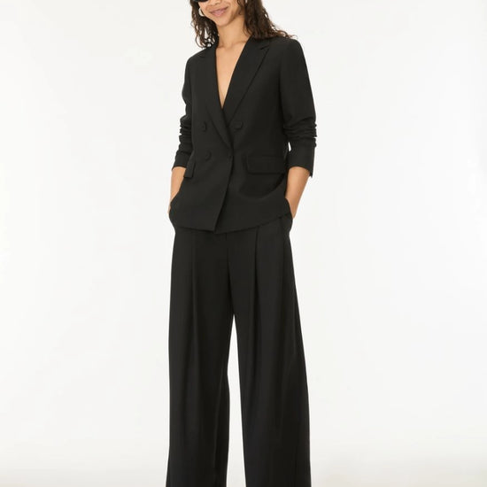 A woman modeling a black tailored blazer with matching wide-leg trousers and black pointed-toe heels against a plain white studio background.
