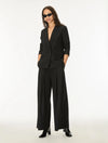 A woman modeling a black tailored blazer with matching wide-leg trousers and black pointed-toe heels against a plain white studio background.