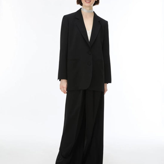 A model wearing a black two-piece suit with a single-breasted blazer and wide-leg trousers, paired with a light-colored choker and silver sandals, against a plain white studio background.
