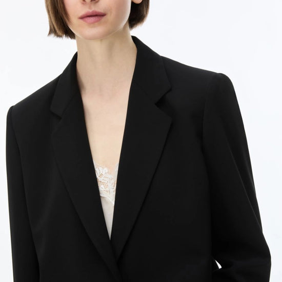 A close-up of a model wearing a black blazer over a white lace top, shot against a plain white background.