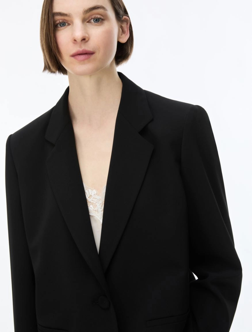 A close-up of a model wearing a black blazer over a white lace top, shot against a plain white background.